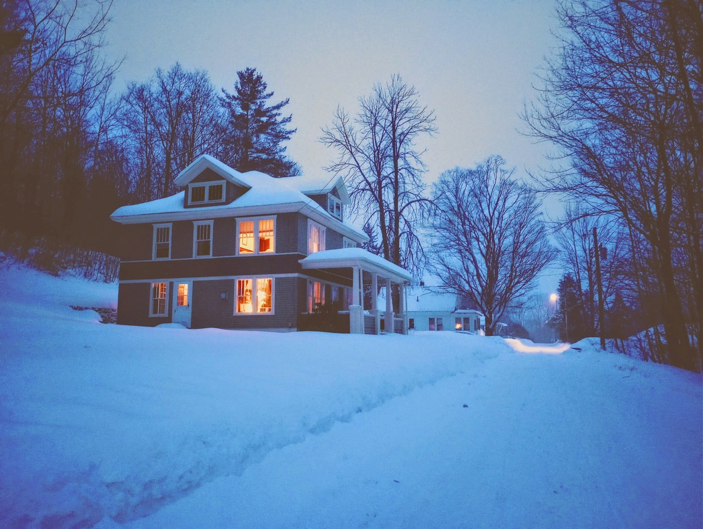 Snow surrounding a home with warmly lit windows at dusk